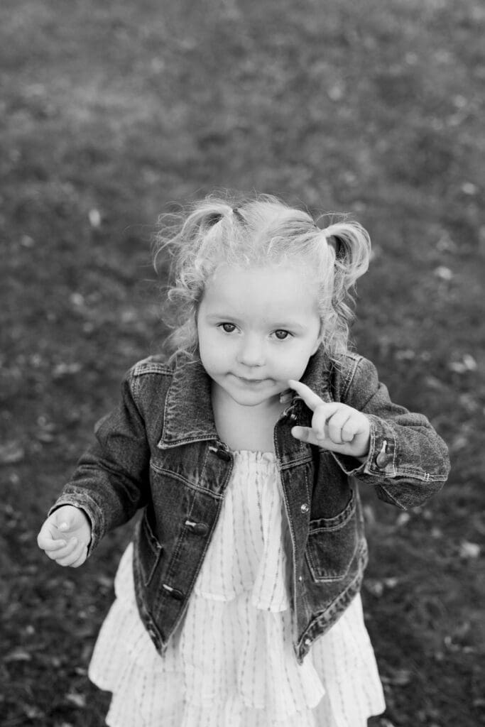 Little girl with pigtails and a denim jacket smiling mischievously with her finger to her lips.