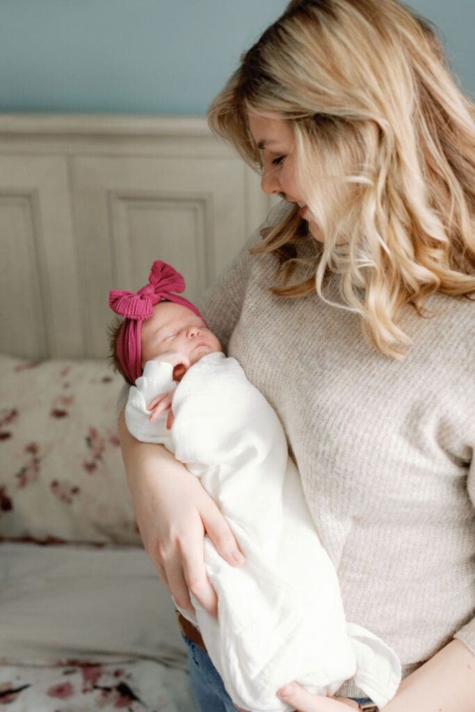 Mother in a beige sweater holding her newborn close while standing next to a bed.