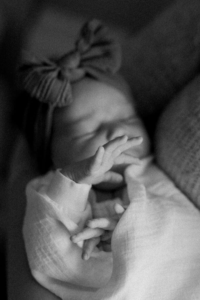 Newborn baby swaddled in white, wearing a bow headband, sleeping peacefully in dim light.