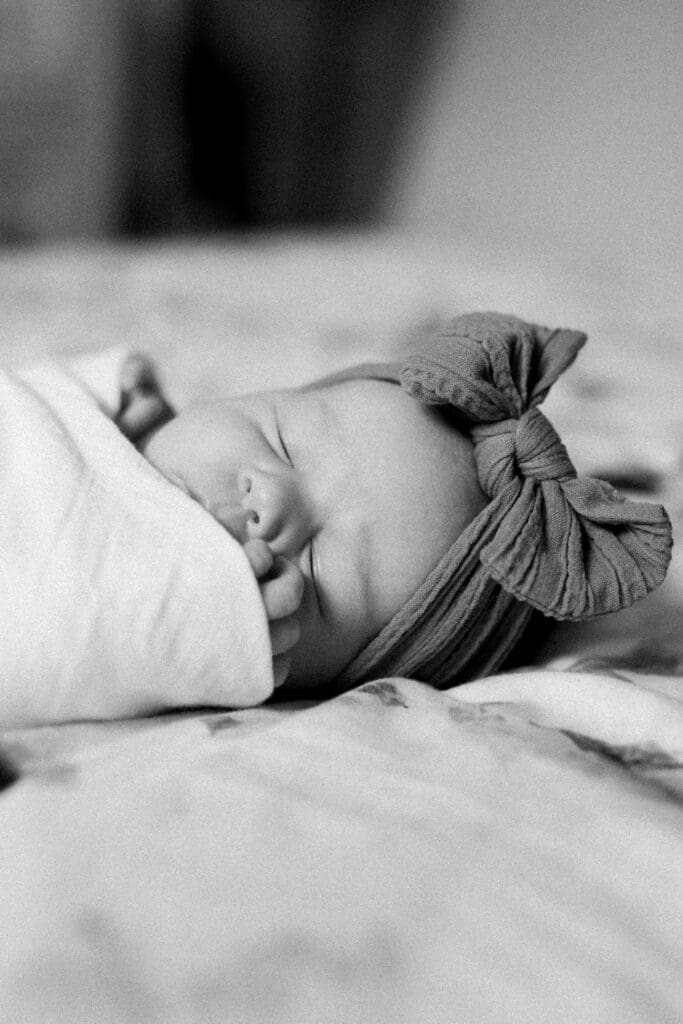 Black-and-white photo of a sleeping newborn wrapped in a soft swaddle with a bow headband.