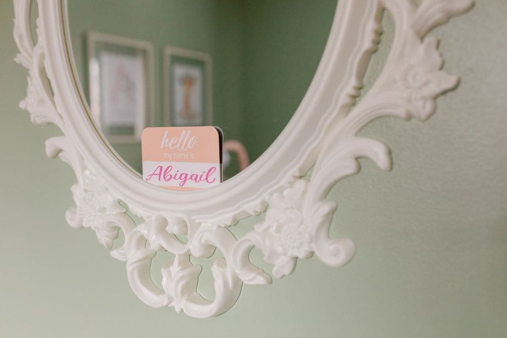 Ornate white mirror with a pink name card reading “Hello Abigail” in a nursery.