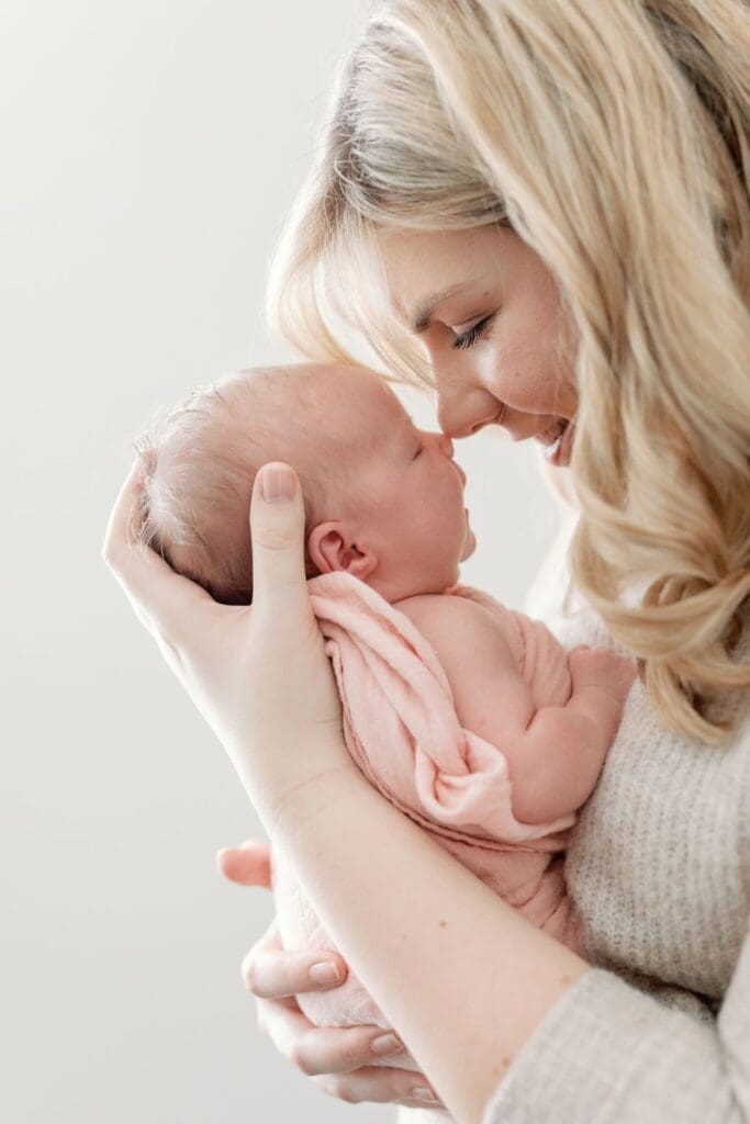 Blonde mother gently touching her nose to her newborn baby’s nose while holding her close.