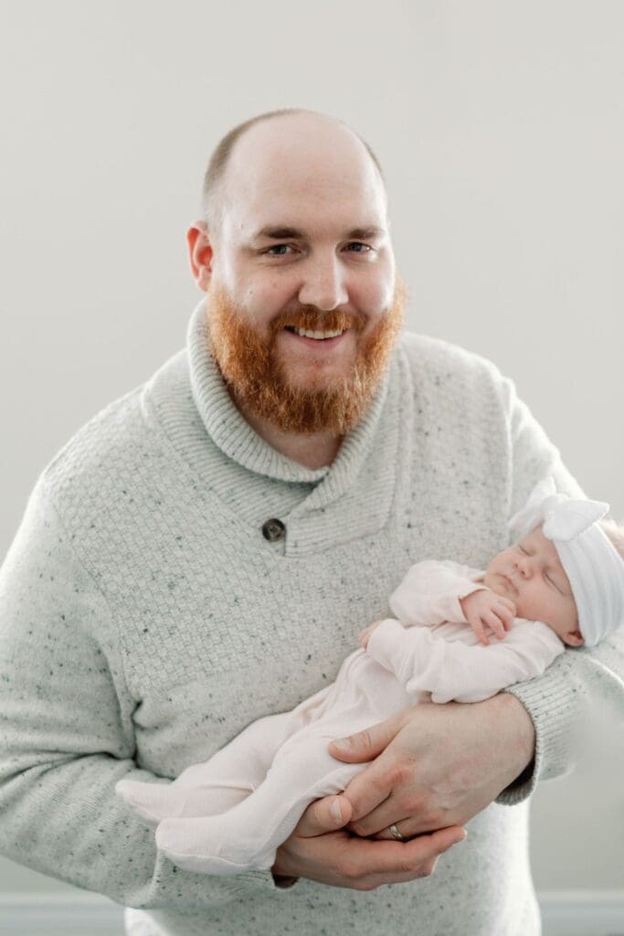 Smiling father in cream sweater holding his newborn daughter in a pink wrap and bow
