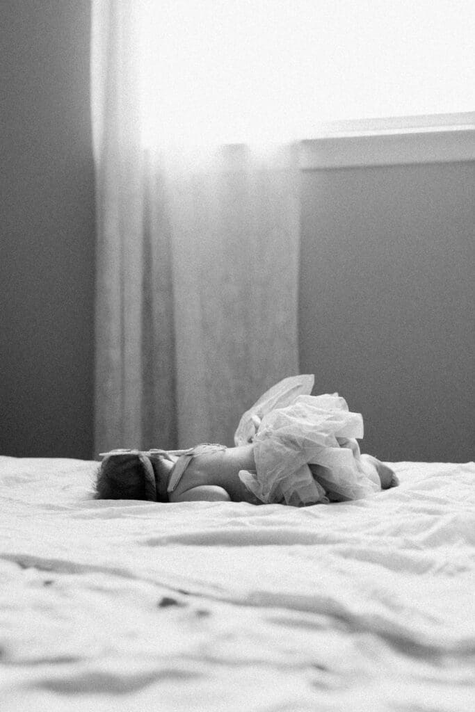 Black and white image of a newborn sleeping on a bed with soft window light illuminating the scene