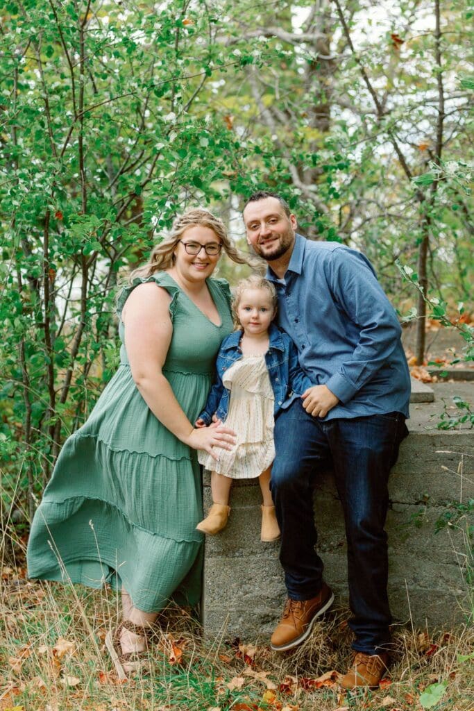 Family of three standing together in a wooded area, all smiling.