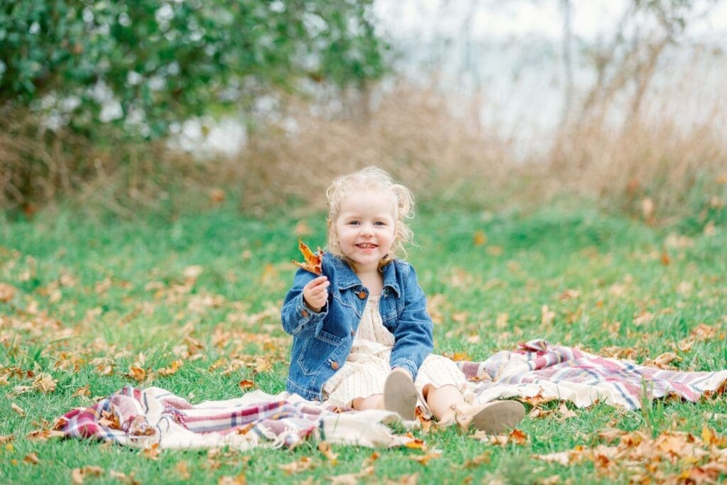 Toddler sitting on the ground holding a leaf, smiling at the camera.