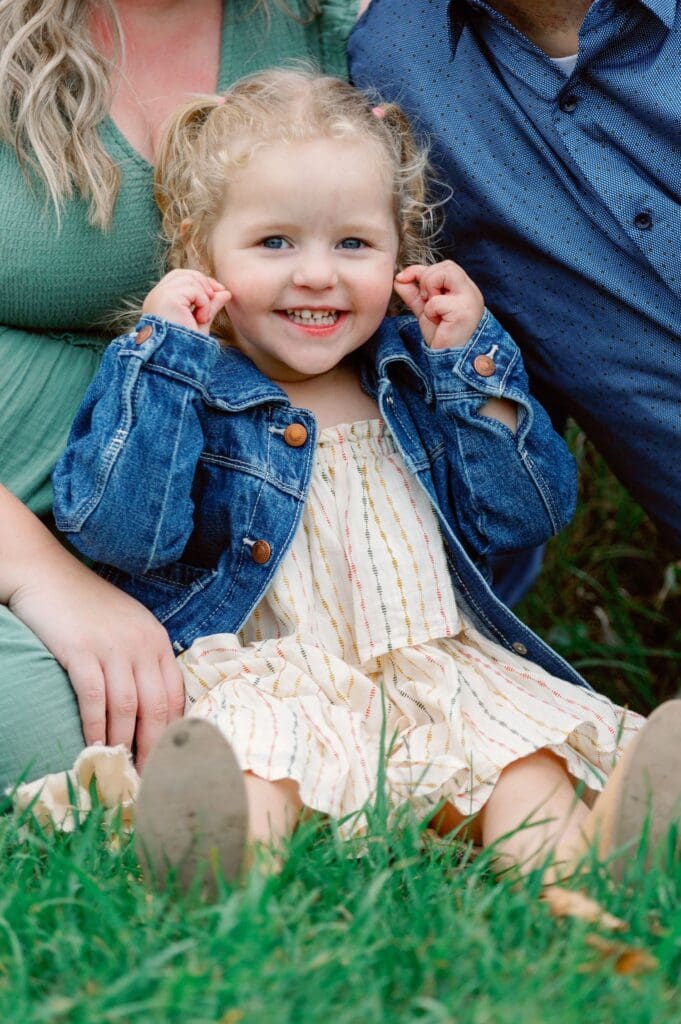 Toddler sitting on the grass in a denim jacket and striped dress, hands at her cheeks and smiling.