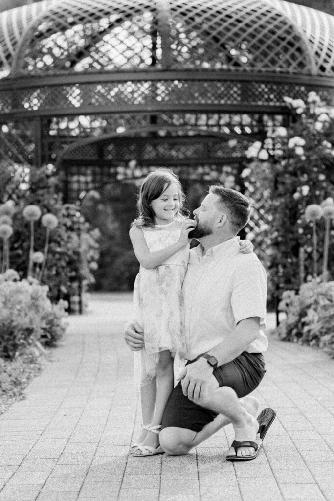 Father smiling at daughter while kneeling in front of a garden gazebo.