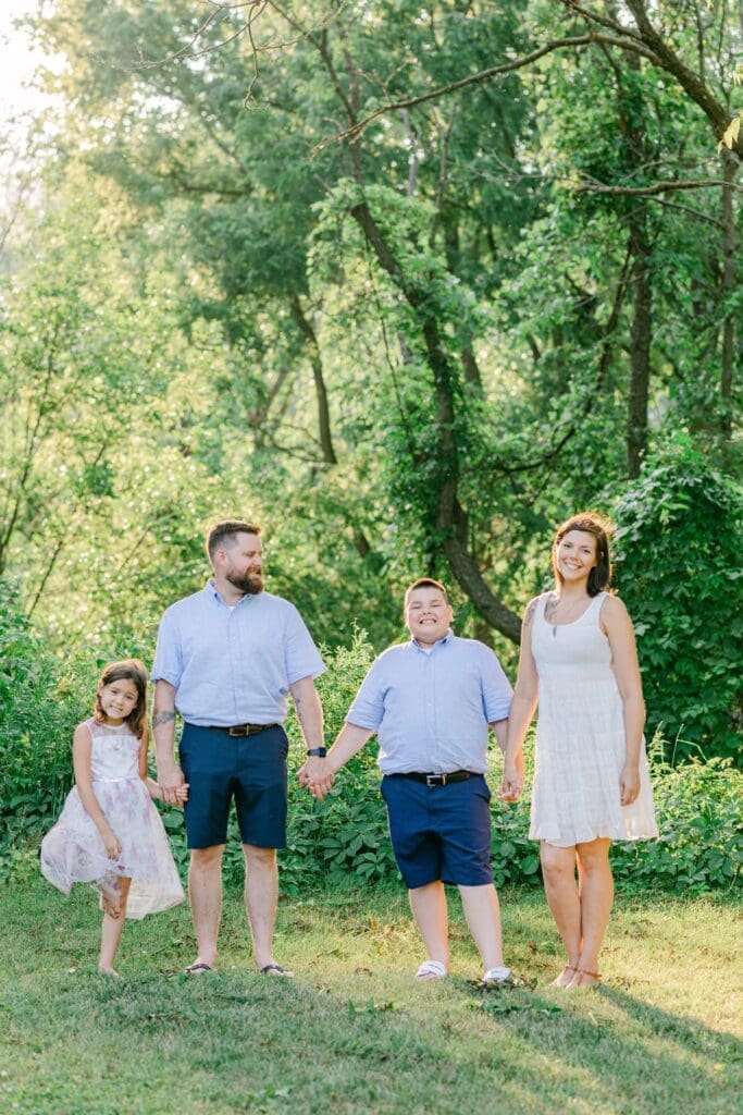 Family of four holding hands and smiling in a sunlit clearing at Burlington Botanical Gardens.