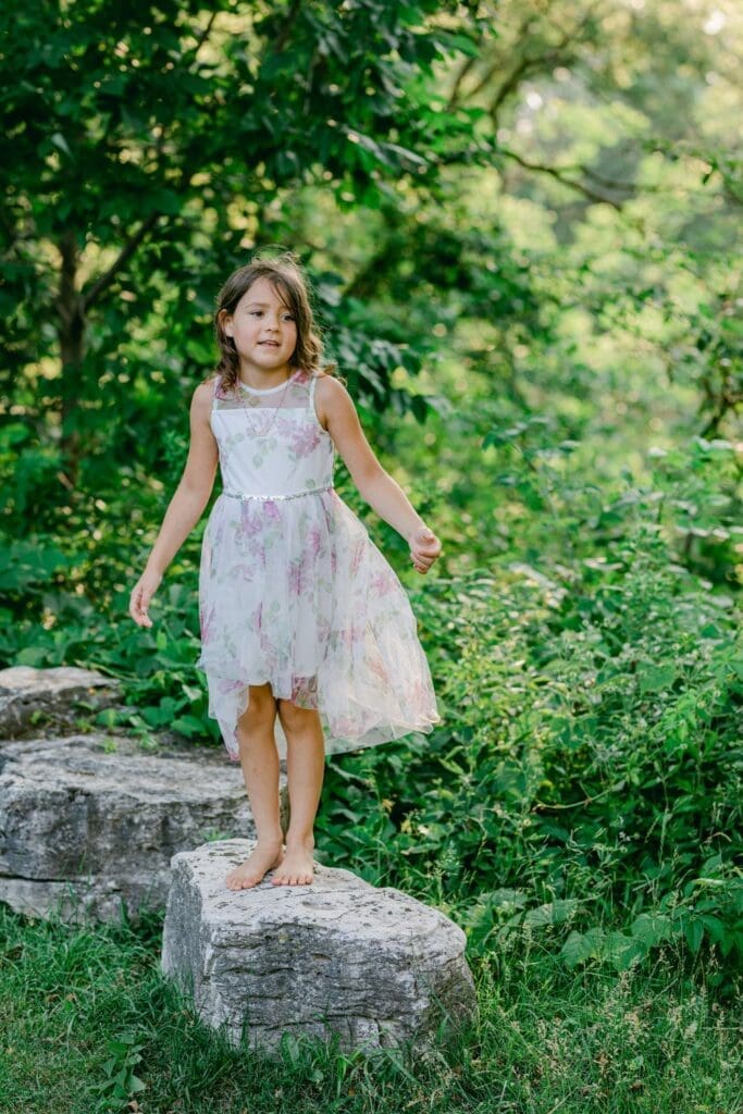  Barefoot girl in floral dress standing on a large garden stone surrounded by greenery.