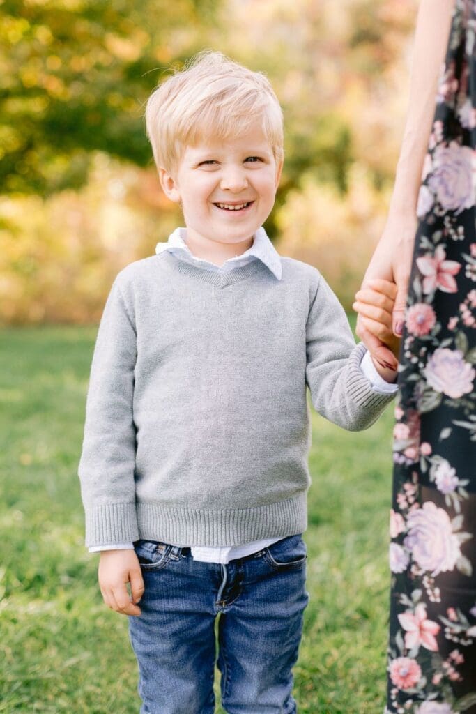  Young blonde boy smiles while holding his mother’s hand during a fall family session