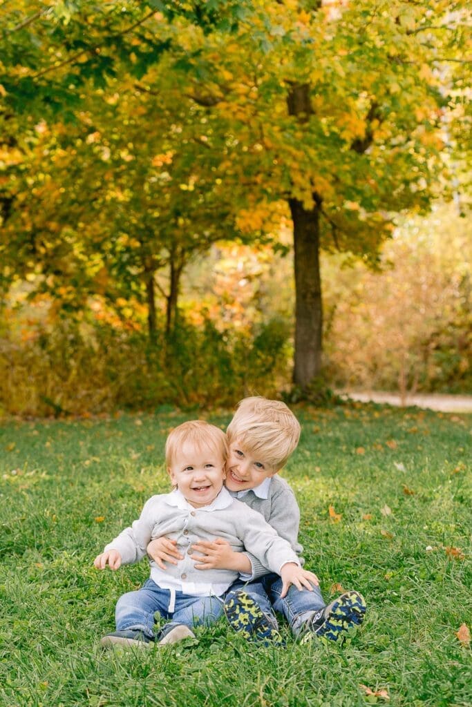 Big brother hugging his younger sibling while sitting on green grass surrounded by autumn leaves