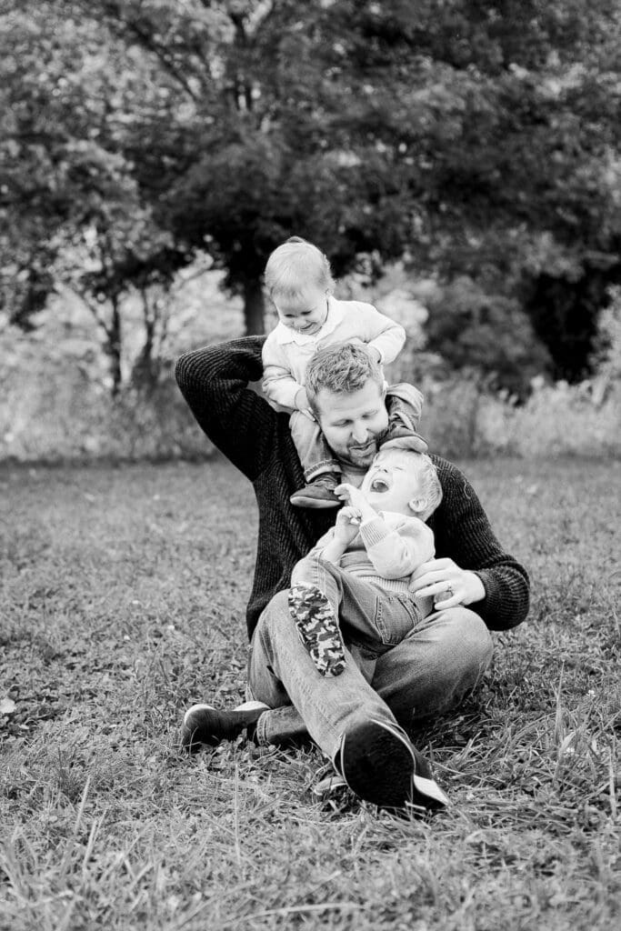 Father sitting on the grass playfully holding his two young children, one on his shoulders and the other in his lap