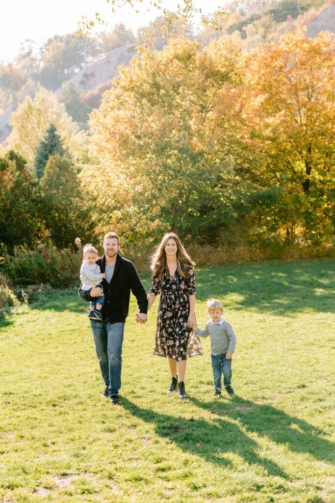 Family of four walking hand-in-hand through a sunlit autumn field with colorful trees in the background.
