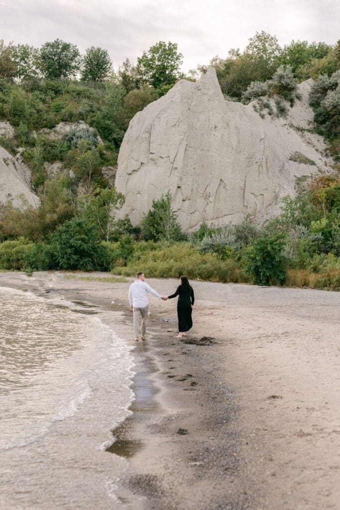 Couple walking hand-in-hand along the sandy shore at Scarborough Bluffs during maternity session
