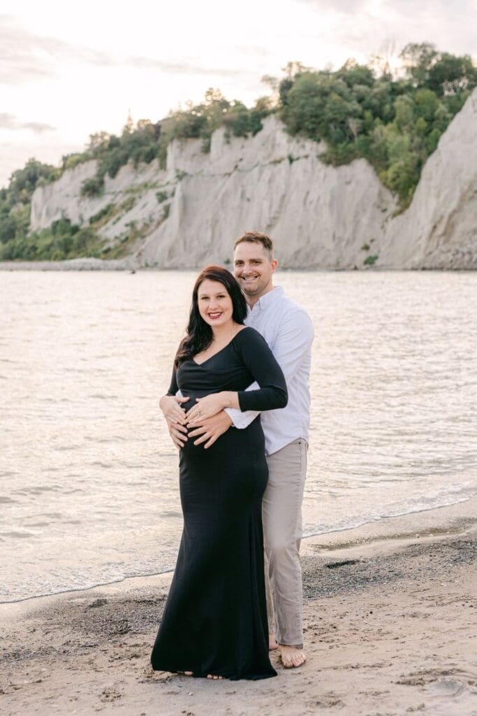 Pregnant woman in black dress embraced by partner by the shore at sunset