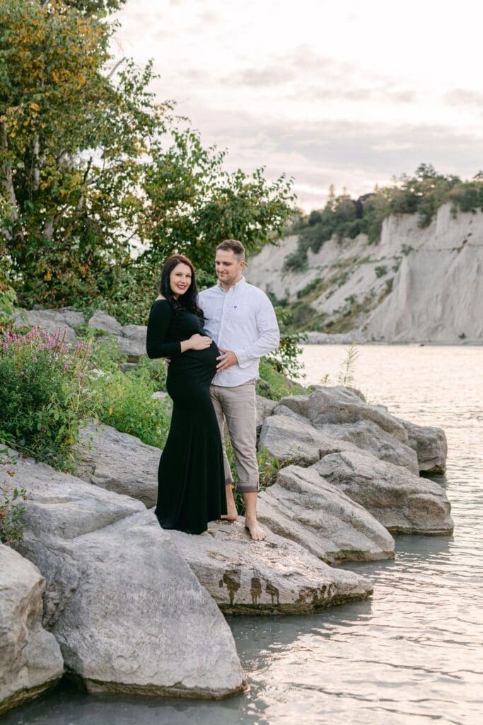 Couple standing barefoot on lakeside rocks during sunset maternity shoot at Scarborough Bluffs