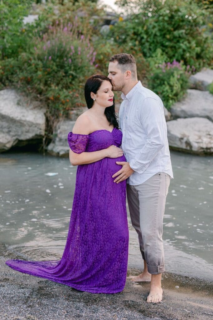 Pregnant woman in flowing purple lace gown stands beside her partner on a beach path