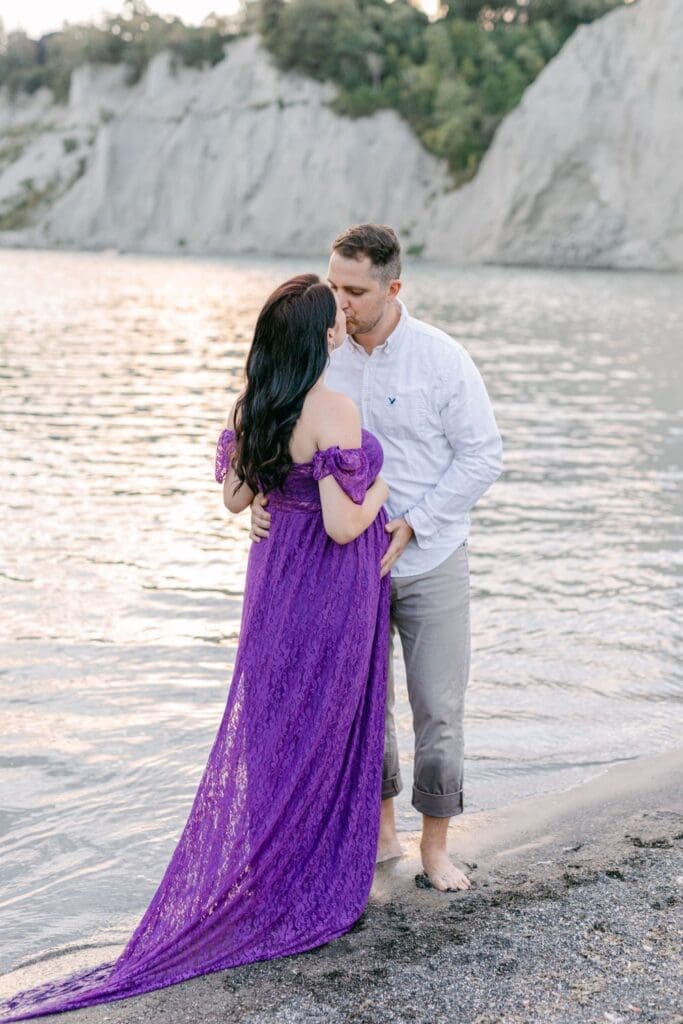 Couple sharing a quiet moment during a maternity session by the lake, woman in purple dress