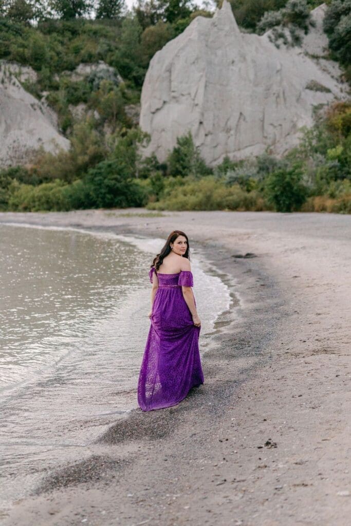 Pregnant woman in a purple lace gown walking along the water's edge at Scarborough Bluffs during sunset
