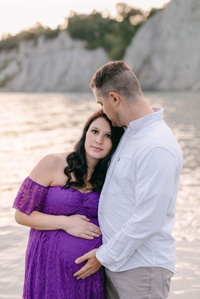 Close-up of couple’s hands resting on pregnant belly with lake in the background