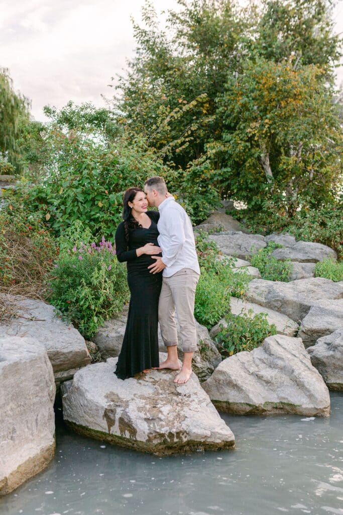 Couple sharing a kiss on large rocks with lake and baby bump visible