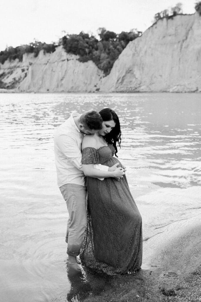Black and white photo of couple embracing at the shoreline during maternity session