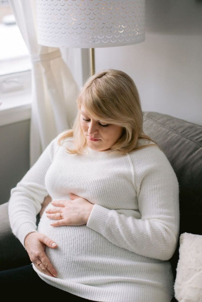 Pregnant woman in a cozy white sweater sitting on a couch and gently holding her belly.