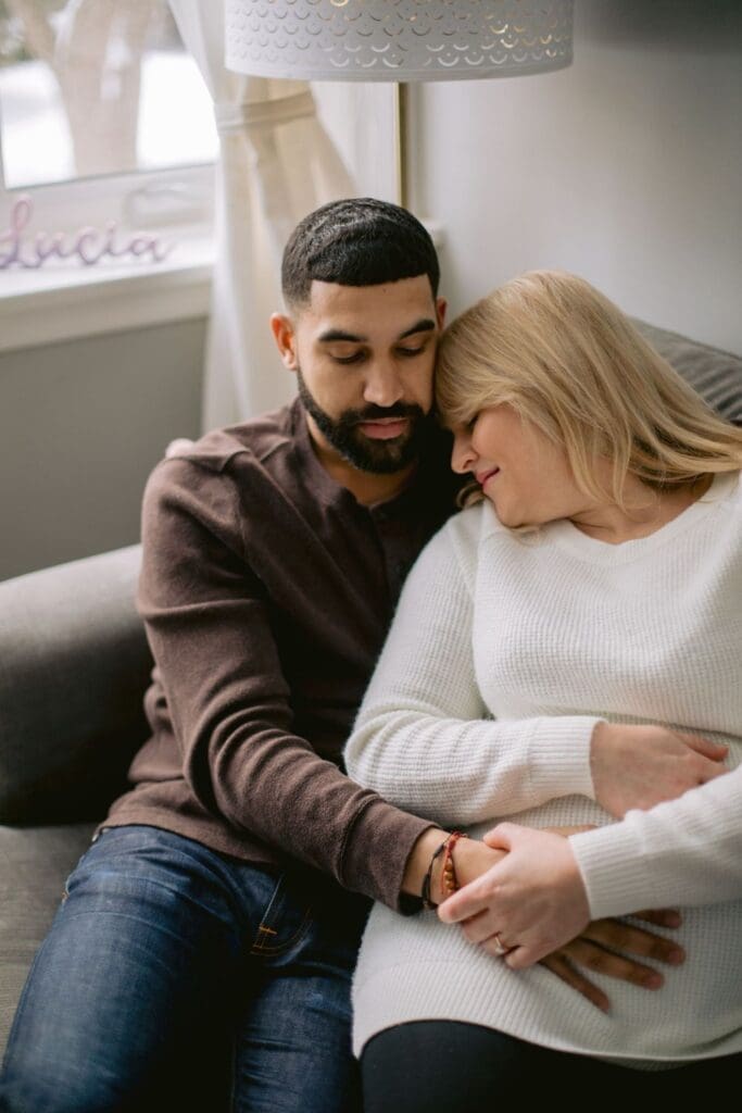 Couple cuddling on the couch, with the mother-to-be resting her head on her partner’s shoulder.