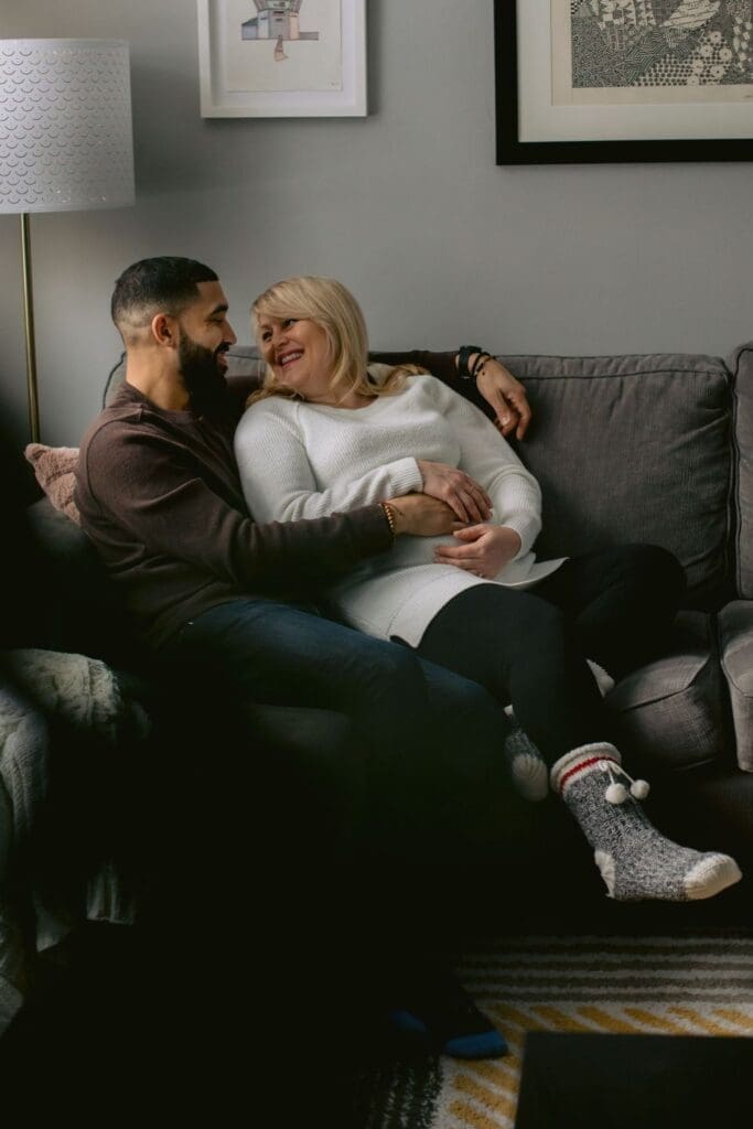 Couple lounging together on a grey couch, smiling at each other during their at-home maternity session.