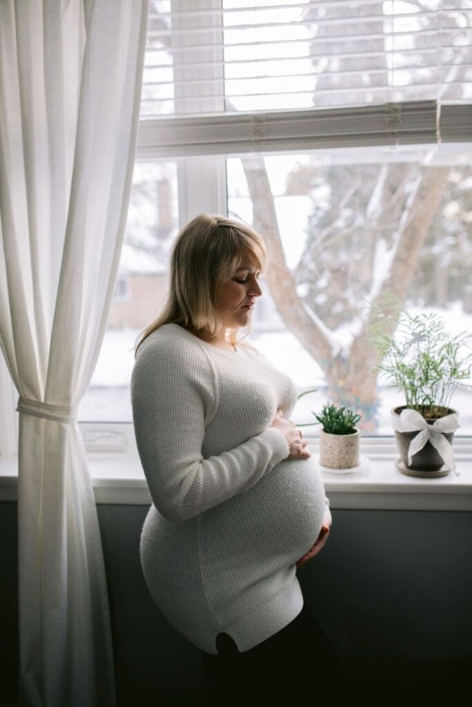 Pregnant woman in a white sweater stands alone by the window, holding her belly and looking down.