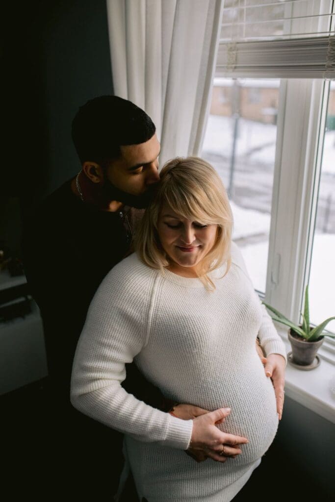 Expecting couple standing by a window, with the father-to-be kissing the mother’s head as she smiles.