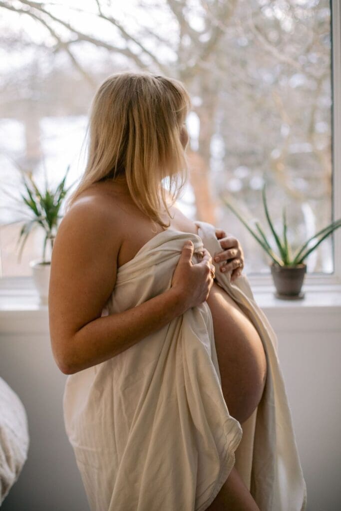 Pregnant woman draped in white fabric stands by a window, gazing outside at a snowy landscape.