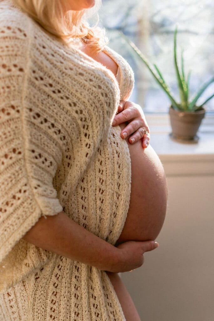Pregnant woman in a cream knit dress holding her belly and standing beside a sunlit window with houseplants.
