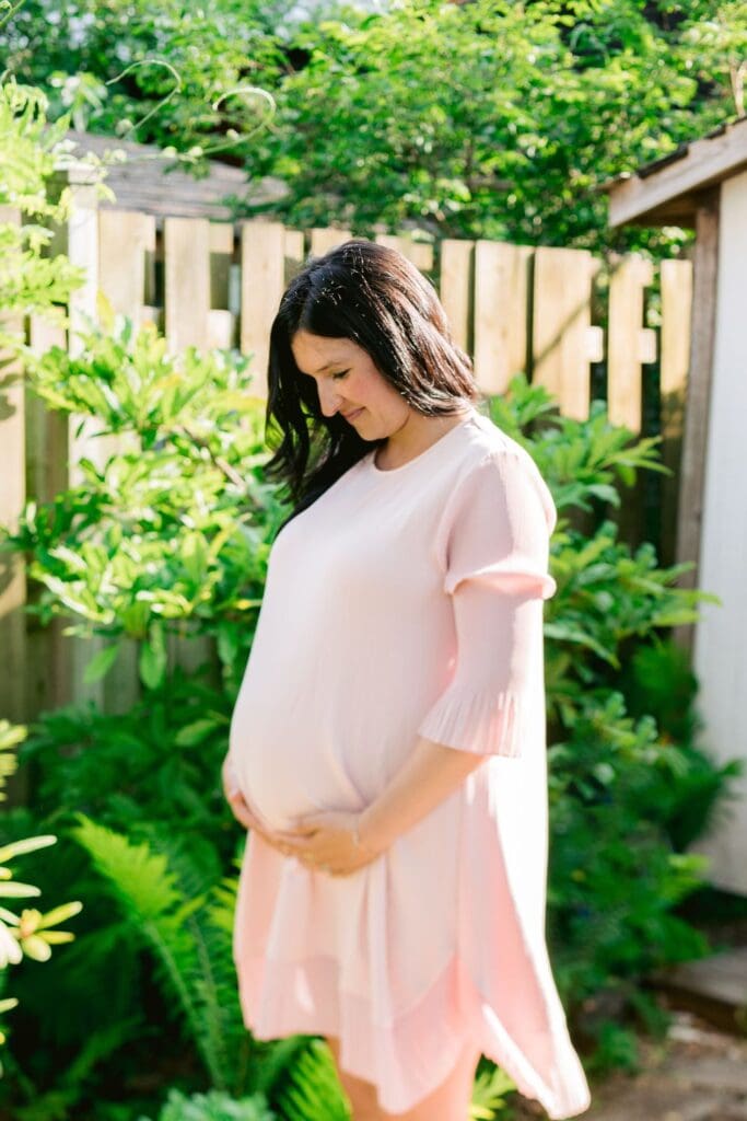 Pregnant woman in a pale pink dress smiling and holding her belly in a lush garden.