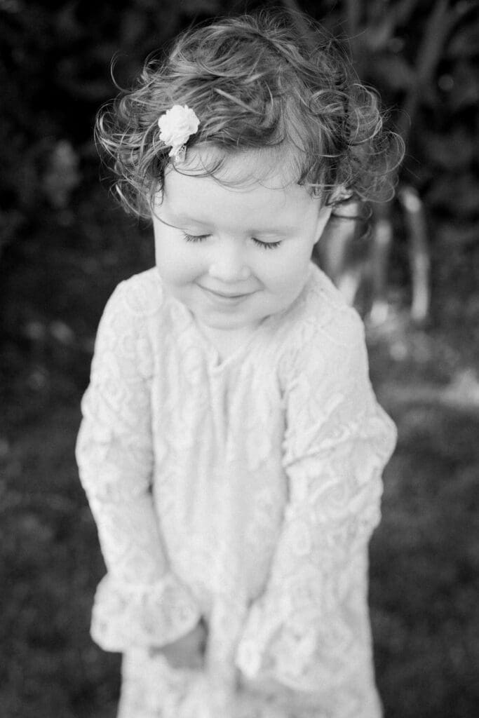 A black and white photo of a young girl in a lace dress looking down shyly with a flower clip in her curly hair.
