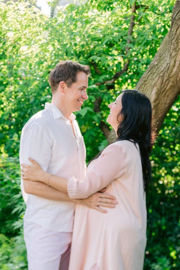 Expecting couple smiling at each other in a green outdoor space.