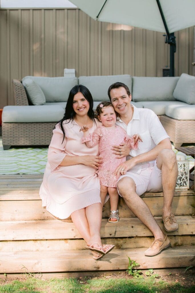 A young family of three sitting on wooden steps with soft smiles and coordinated pastel outfits.