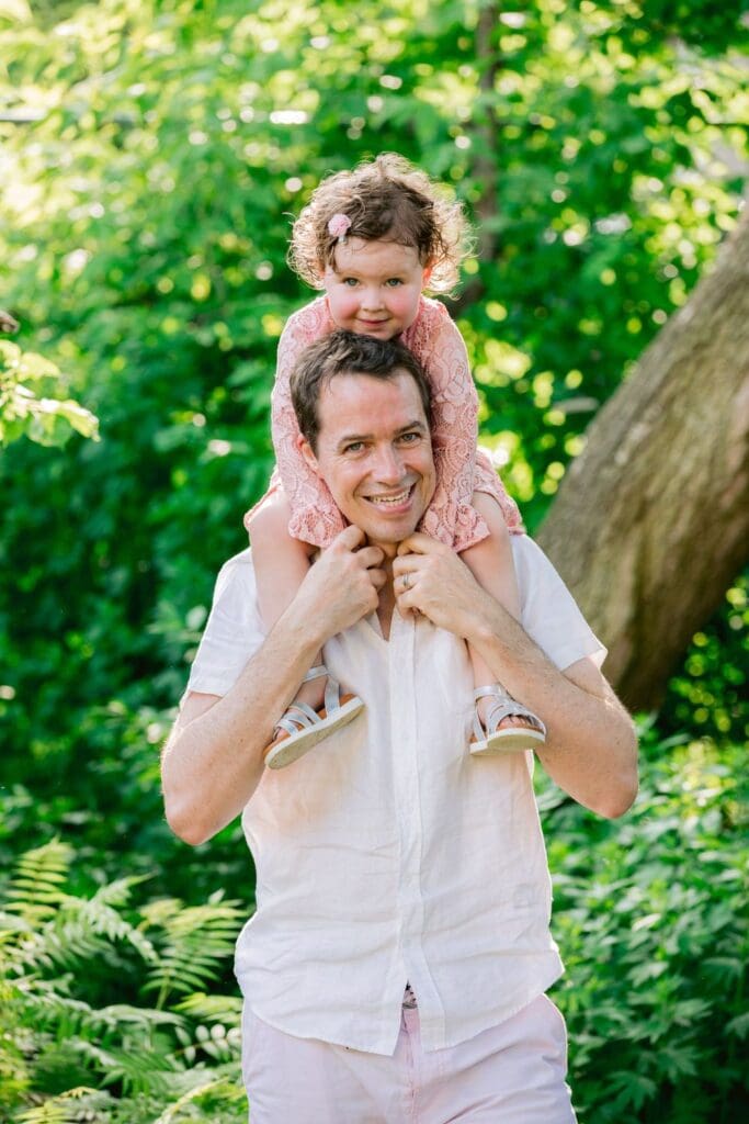 Father carrying daughter on his shoulders while both smile outdoors.