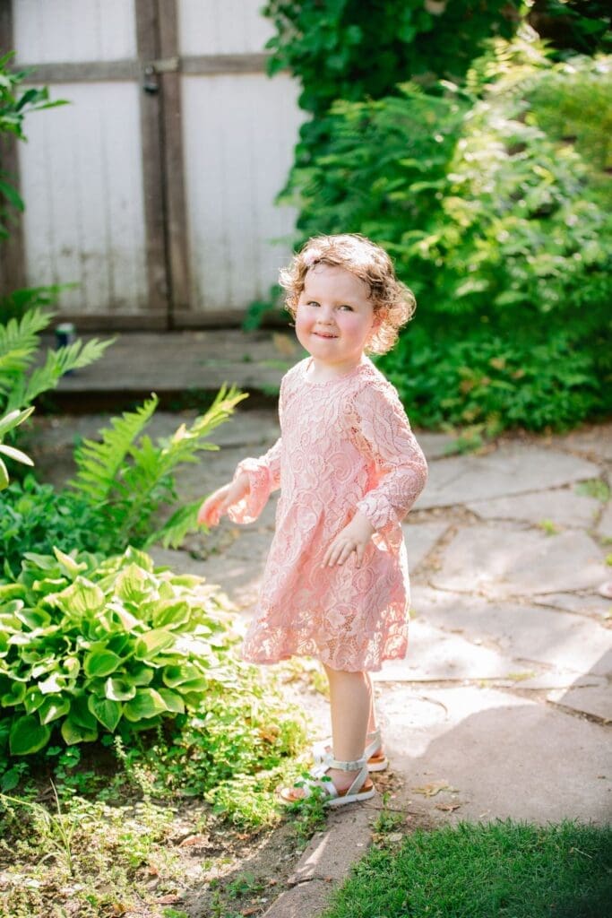 A little girl in a pink lace dress smiling shyly while walking along a garden path.