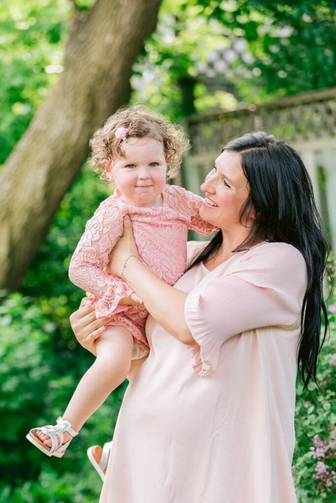A pregnant woman in a blush pink dress holds her toddler daughter, both smiling, surrounded by greenery.
