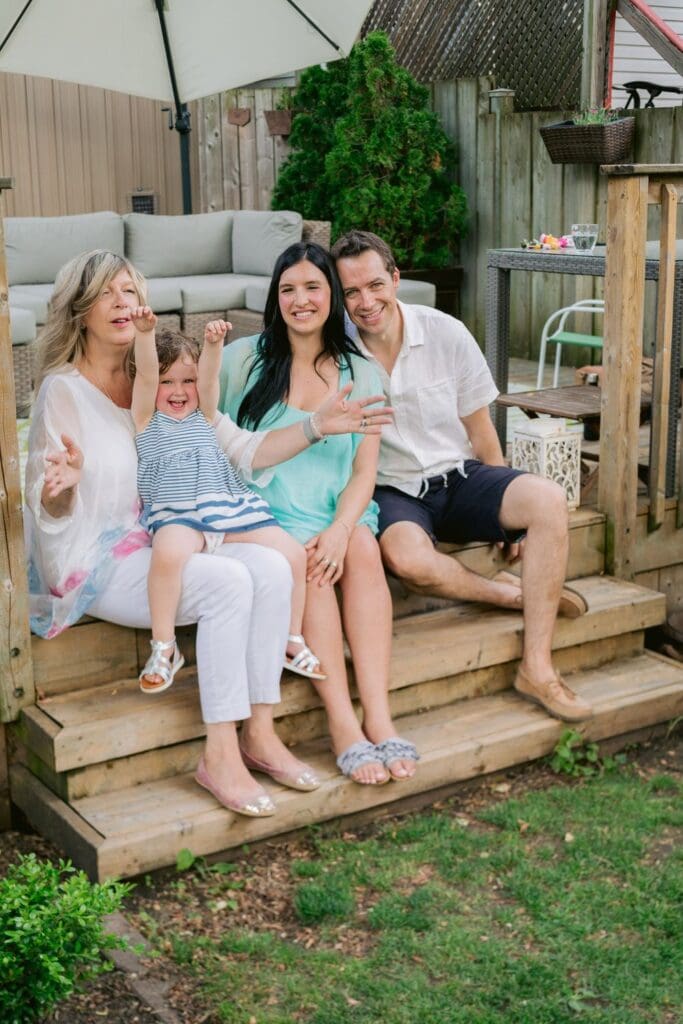 A grandmother, young family, and toddler sitting on wooden steps in a backyard, smiling and enjoying time together.