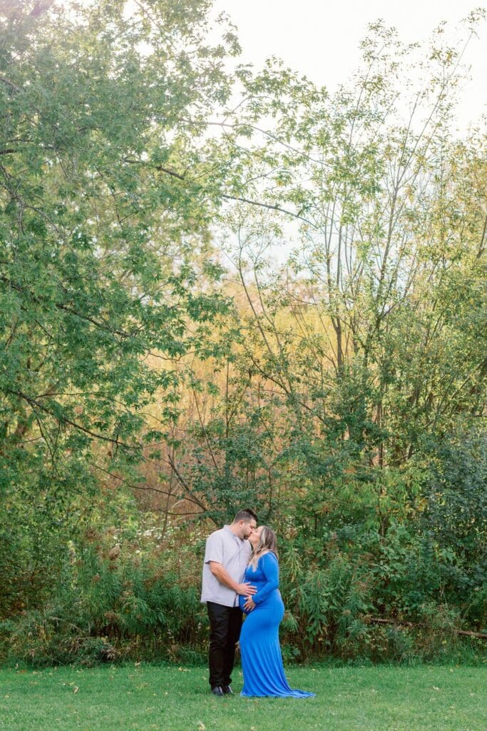A couple shares a kiss while standing in a grassy field, the woman in a flowing blue maternity dress.