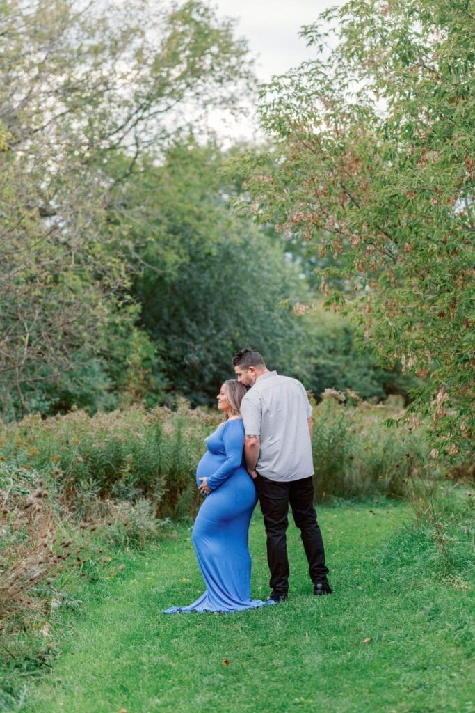 Pregnant woman in a blue gown and her partner sharing a quiet moment on a nature trail.