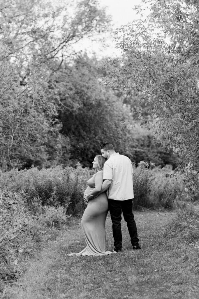 Black and white photo of couple expecting baby, standing on grassy trail with trees around them.