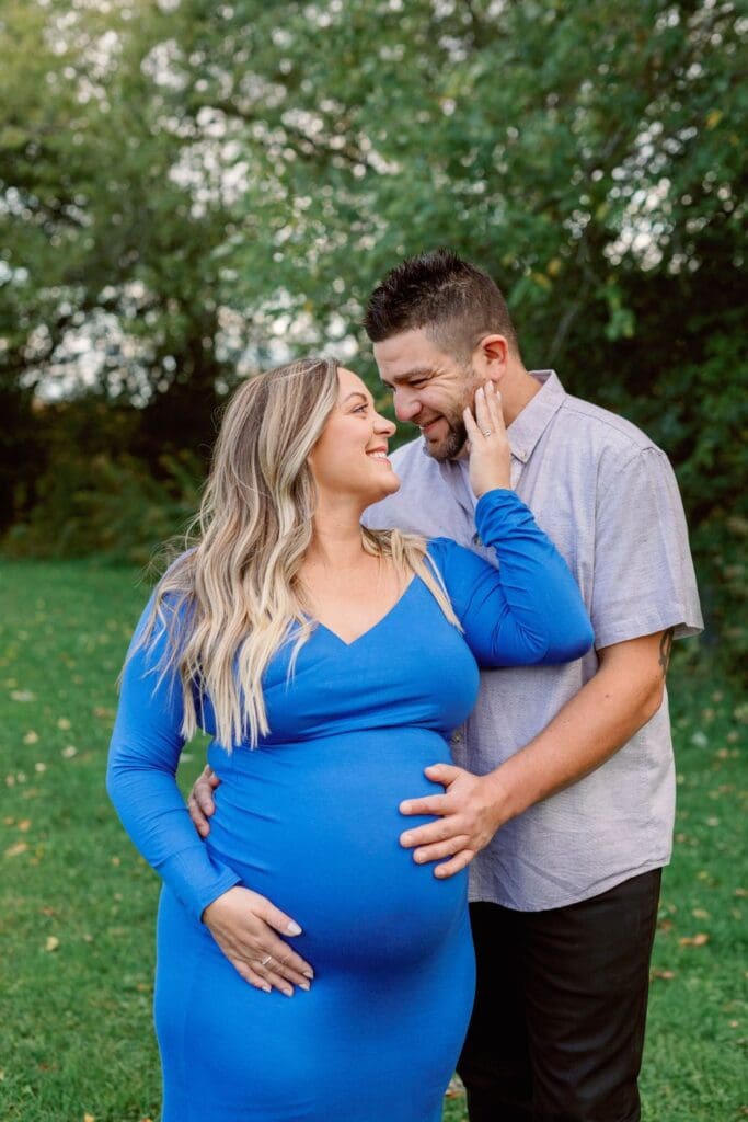 A woman in a blue maternity dress smiles up at her partner as she lovingly touches his face.