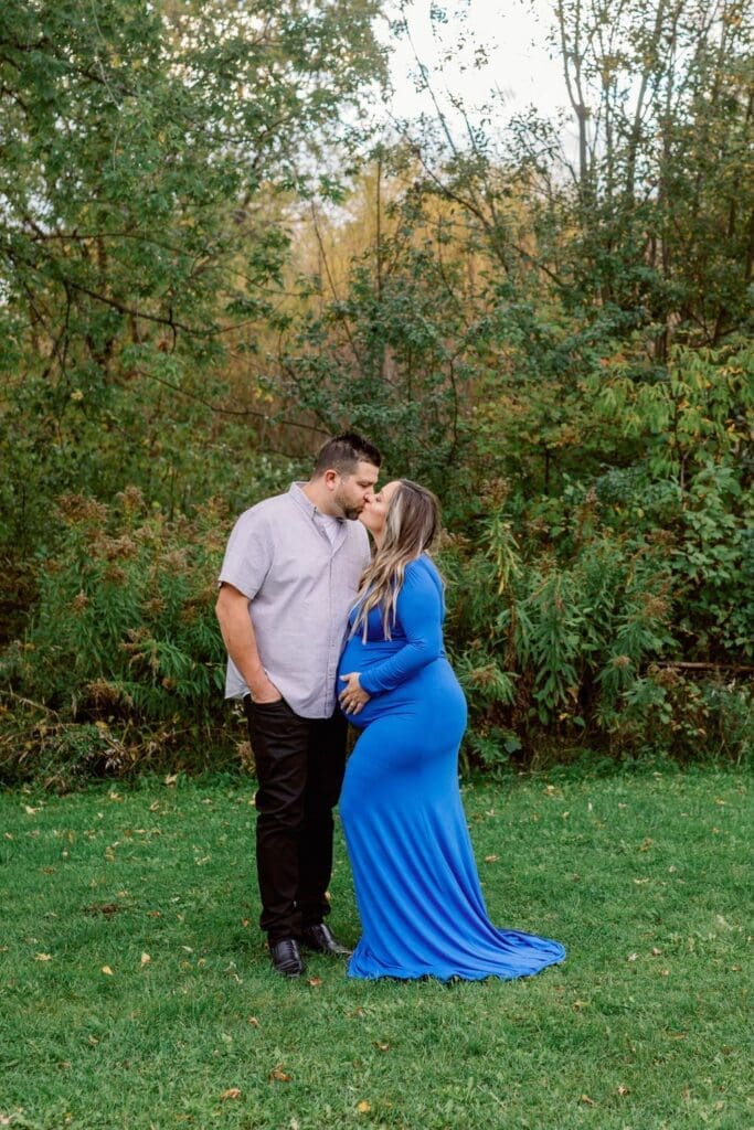 Expecting couple sharing a kiss outdoors, woman in a long blue maternity gown.