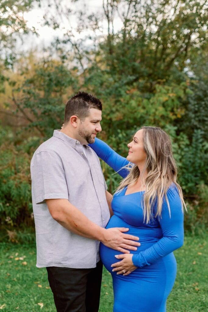 A pregnant woman in a royal blue dress cradles her belly as she gazes at her partner, who holds her gently.