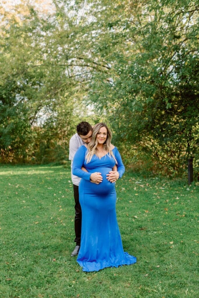 A man stands behind his pregnant partner, wrapping his arms around her belly in a green park setting.