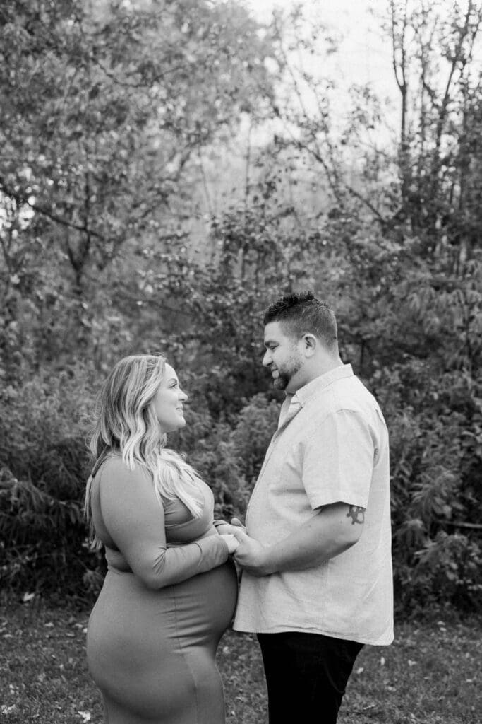 Black-and-white photo of a couple standing face to face, holding hands and smiling at each other in a forested area.
